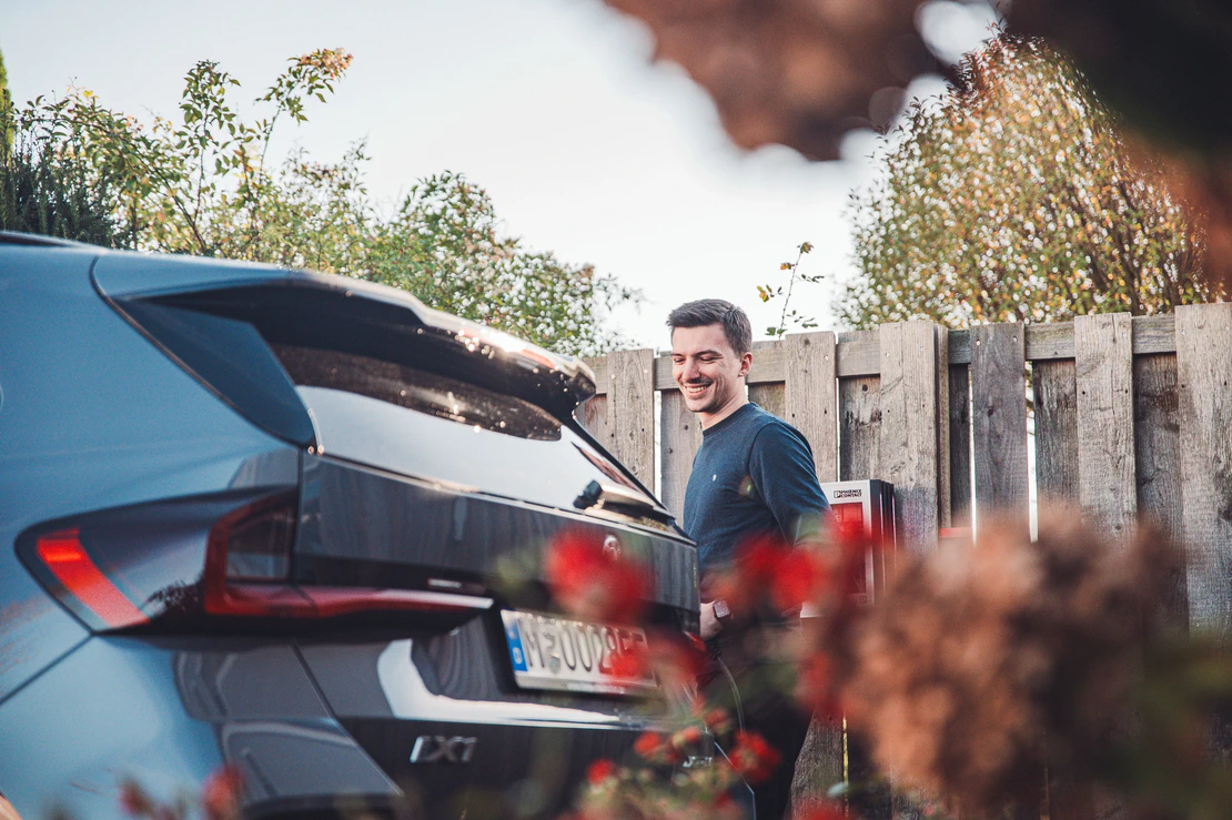 Eine Person steht neben dem Heck eines grauen Autos mit europäischem Kennzeichen. Im Hintergrund sind ein Holzzaun und etwas Grün zu sehen, im Vordergrund unscharfe Blumen. 