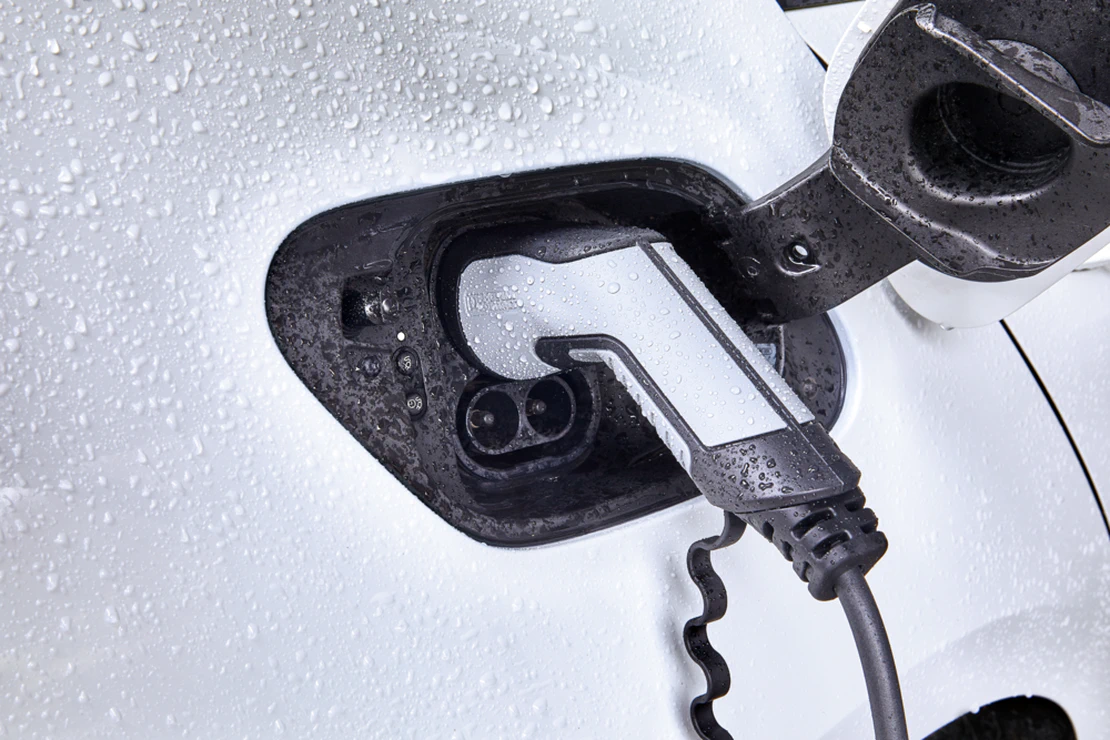 Nahaufnahme eines Elektroautos beim Ladevorgang: Ein Ladekabel ist in den Ladeanschluss eines weißen Fahrzeugs eingesteckt. Der Ladebereich ist geöffnet und mit Wassertropfen bedeckt, was auf den Einsatz im Außenbereich hinweist.